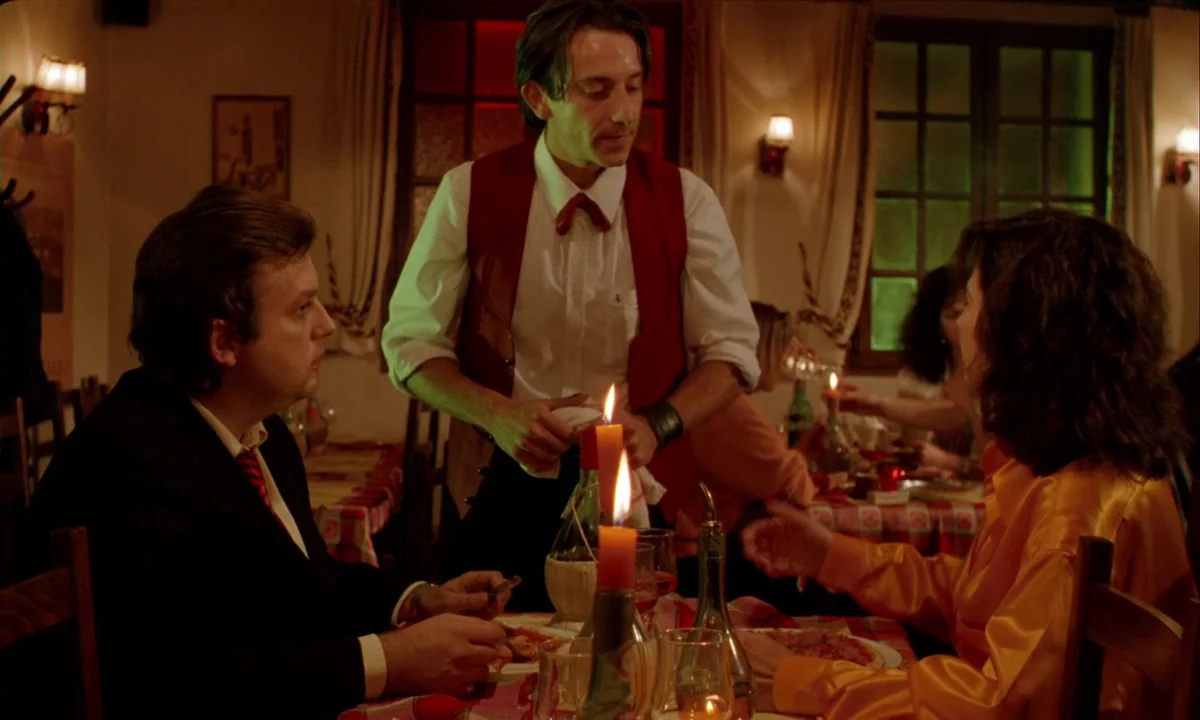 Betty Blue (1986) movie still: medium — A dimly lit restaurant features checkered tablecloths and wooden chairs. A waiter stands between a seated…