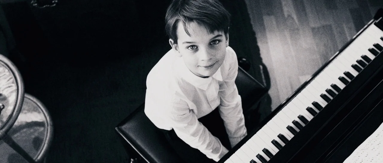 Sound of Noise (2010) movie still: medium — A high-angle view shows a young boy sitting on a black bench. He sits positioned in front of a piano…