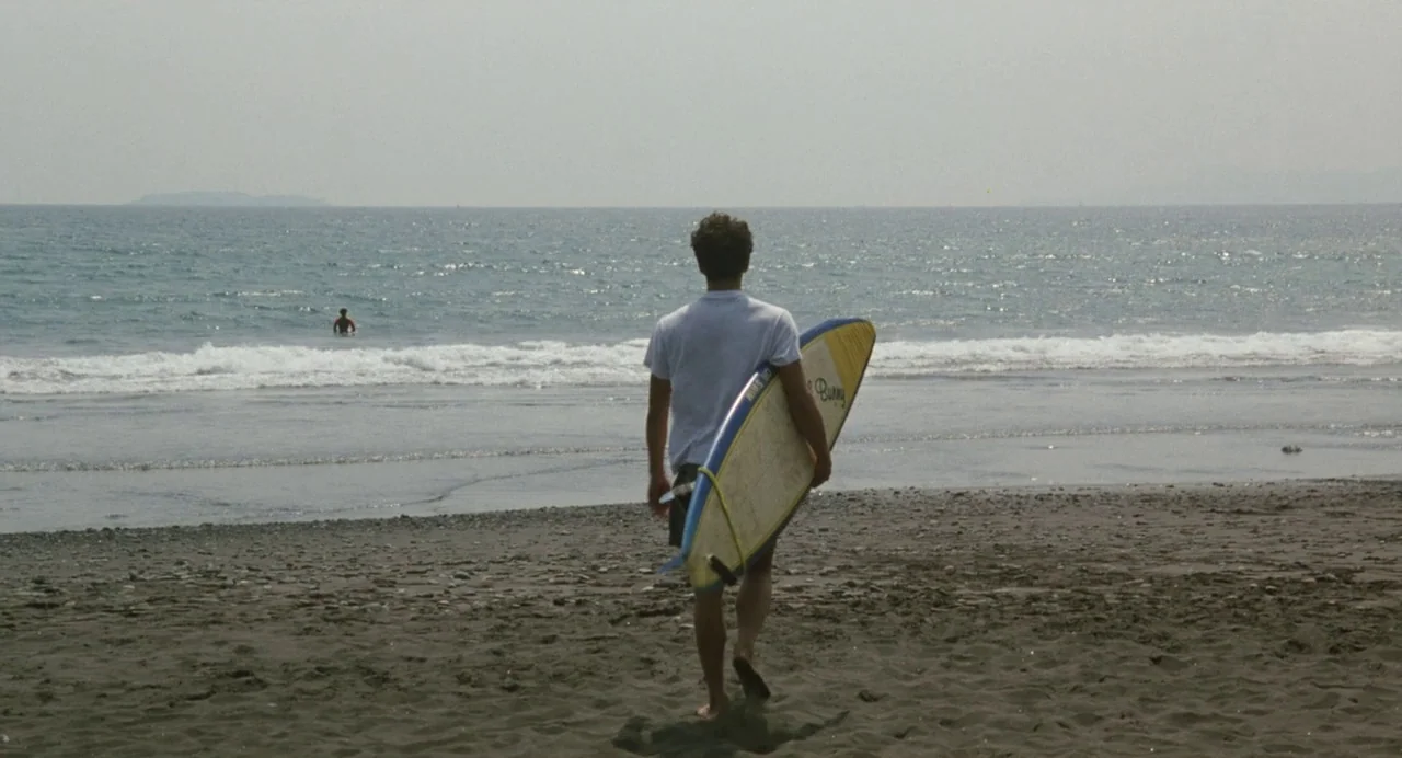 A Scene at the Sea (1991) movie still: fullbody — A dark sand beach meets the calm ocean under an overcast sky. A young man carries a yellow and…