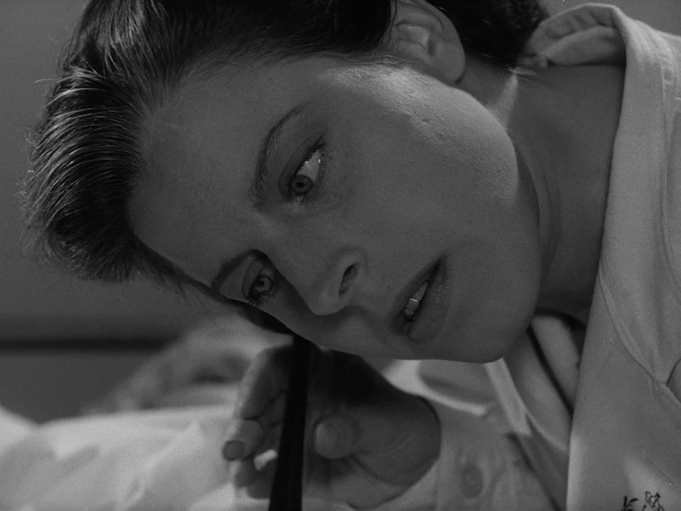 Brink of Life (1958) movie still: closeup — A close-up view of a woman lying in bed inside a bedroom. She holds a telephone receiver to her ear…