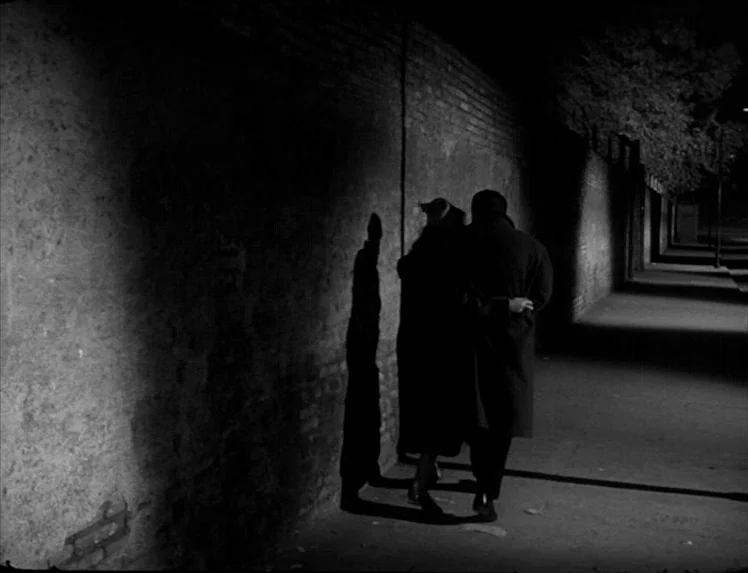 I Vitelloni (1953) movie still: fullbody — A man and a woman walk alongside a dark brick wall at night. Their silhouettes are cast against the…