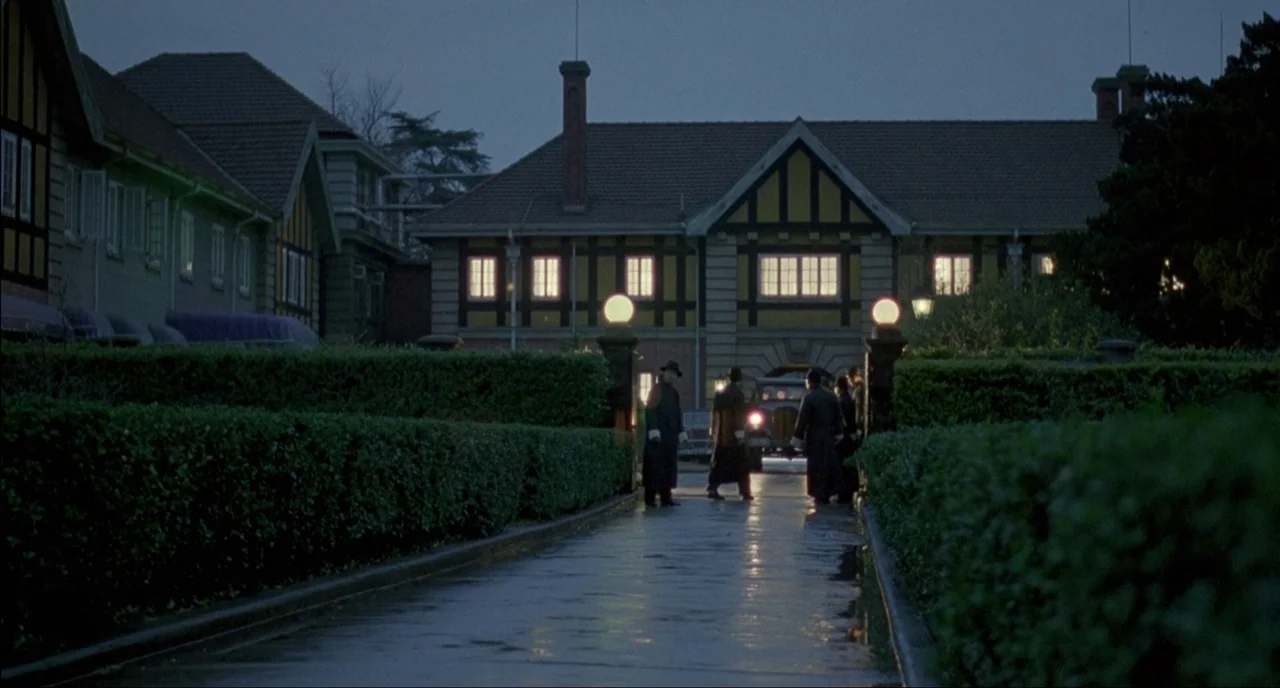 Shanghai Triad (1995) movie still: wide — A dark, wet driveway leads to a large, half-timbered building with illuminated windows. Several men in…