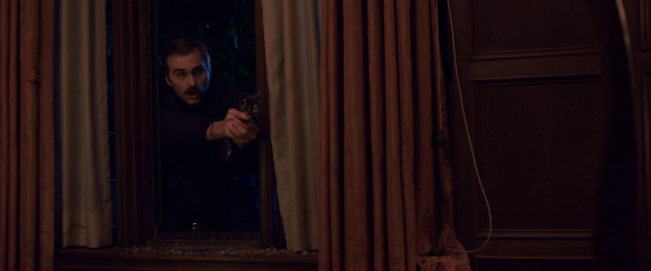 You’re Next (2013) movie still: medium — A dark, wood-paneled room interior viewed through a window frame with heavy curtains. A police officer aims…