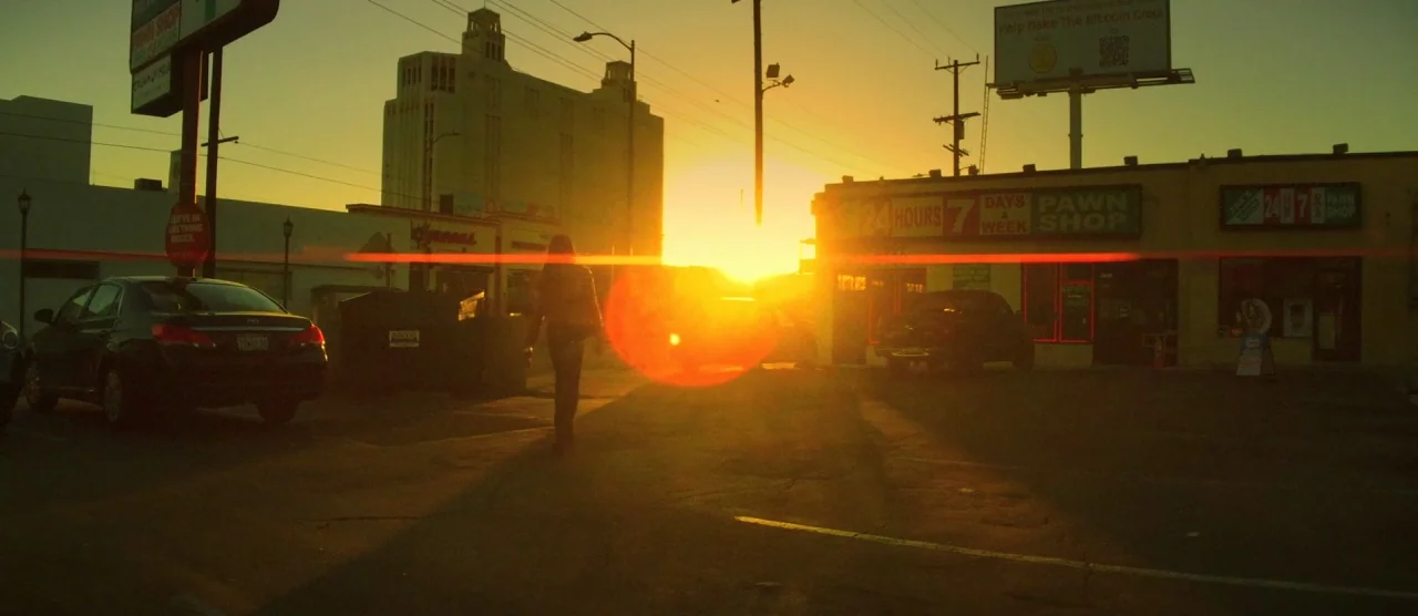 Tangerine (2015) movie still: wide — A desolate urban parking lot at sunset features a pawn shop and industrial buildings. A person walks slowly…