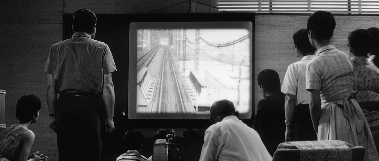 High and Low (1963) movie still: wide — A group of people watch a projected film in a minimalist, wood-paneled room. The projection shows a…
