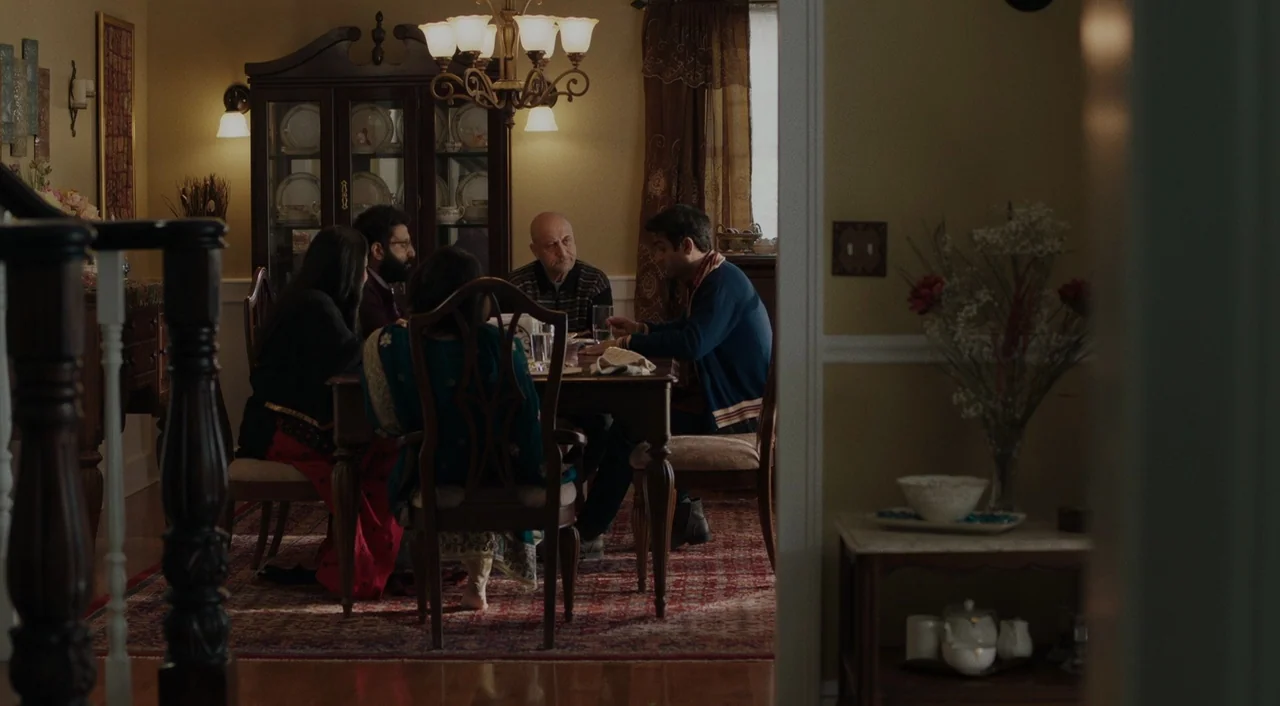 The Big Sick (2017) movie still: medium — A dimly lit room contains a dark wood dining table set for a meal. Four adults sit around the table having…