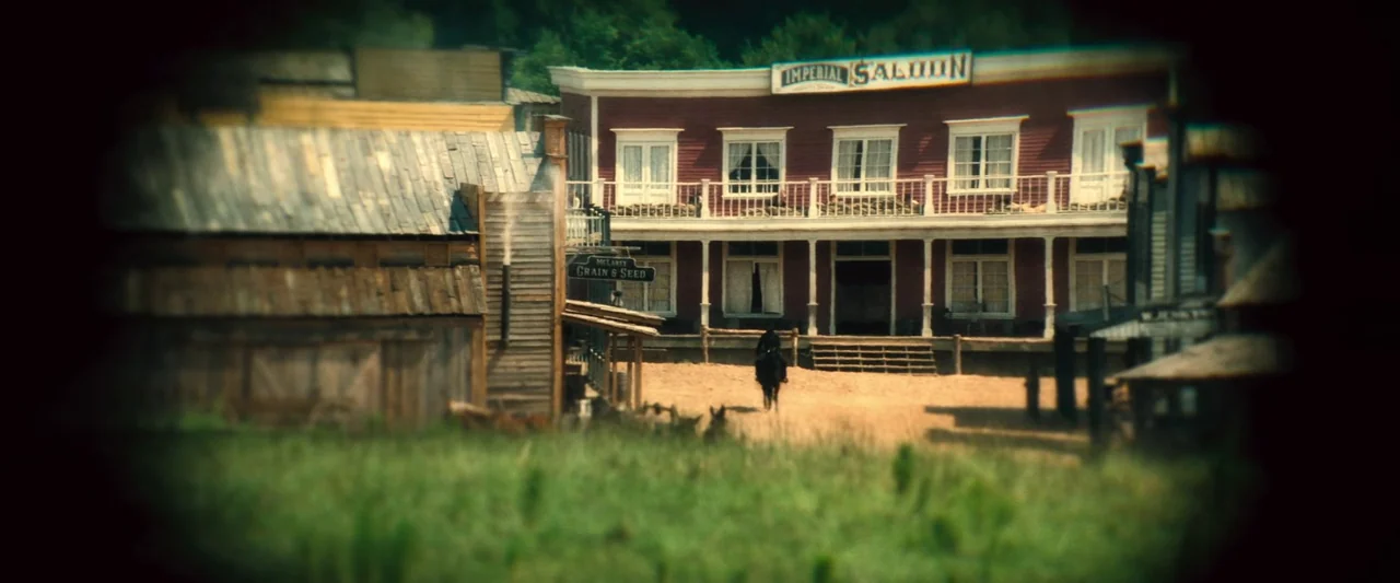 The Magnificent Seven (2016) (2016) movie still: establishing — A wooden old west town square features a prominent red saloon building with a…