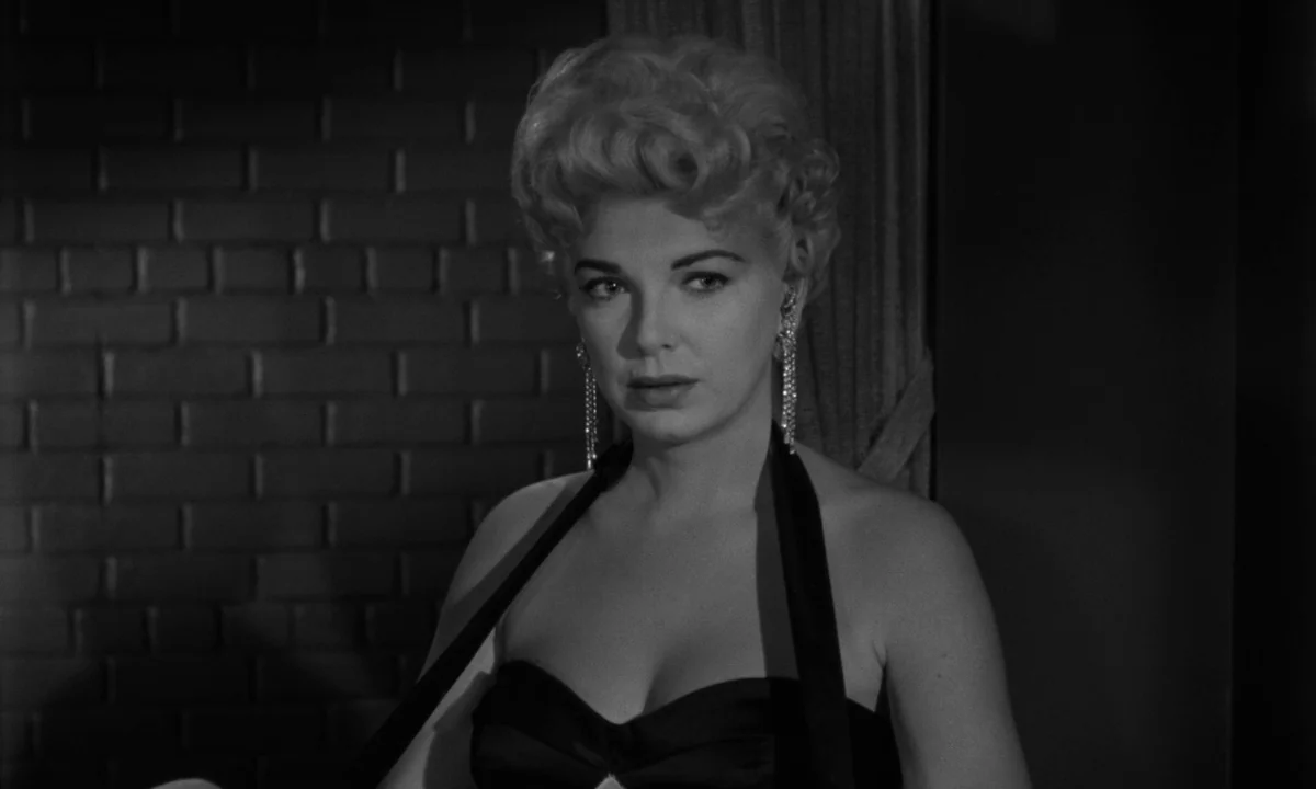 Sweet Smell of Success (1957) movie still: medium — A dark indoor space features a wall with a brick texture in the background. A woman in a dark…