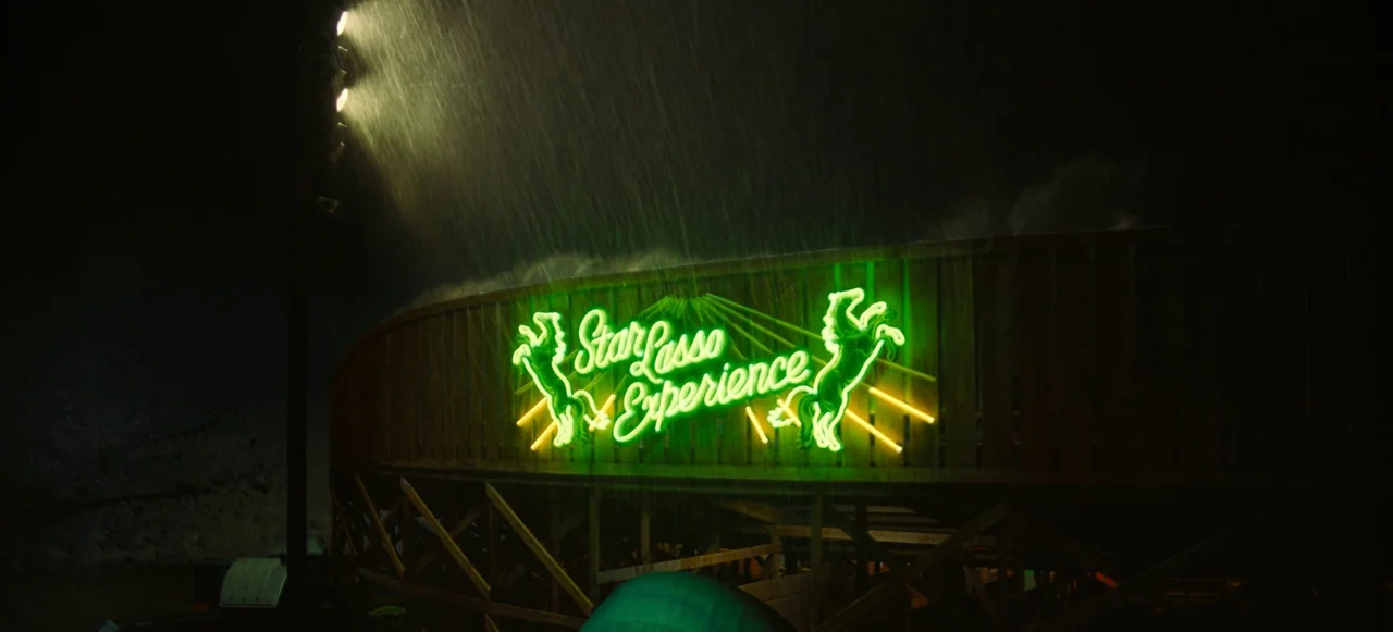 Nope (2022) movie still: wide — A wooden structure features a glowing neon sign in a dark, outdoor setting at night. Heavy rain pours down, catching…