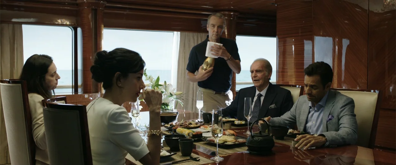 Overboard (2018) movie still: medium — A wood-paneled dining room inside a yacht with windows overlooking the ocean. Four people are seated at a…