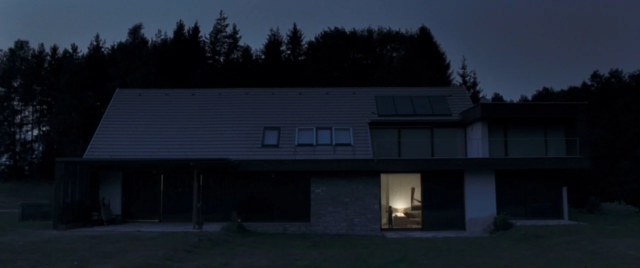 Goodnight Mommy (2014) movie still: wide — A modern two-story home stands at the edge of a dense dark forest at night. A single interior light glows…