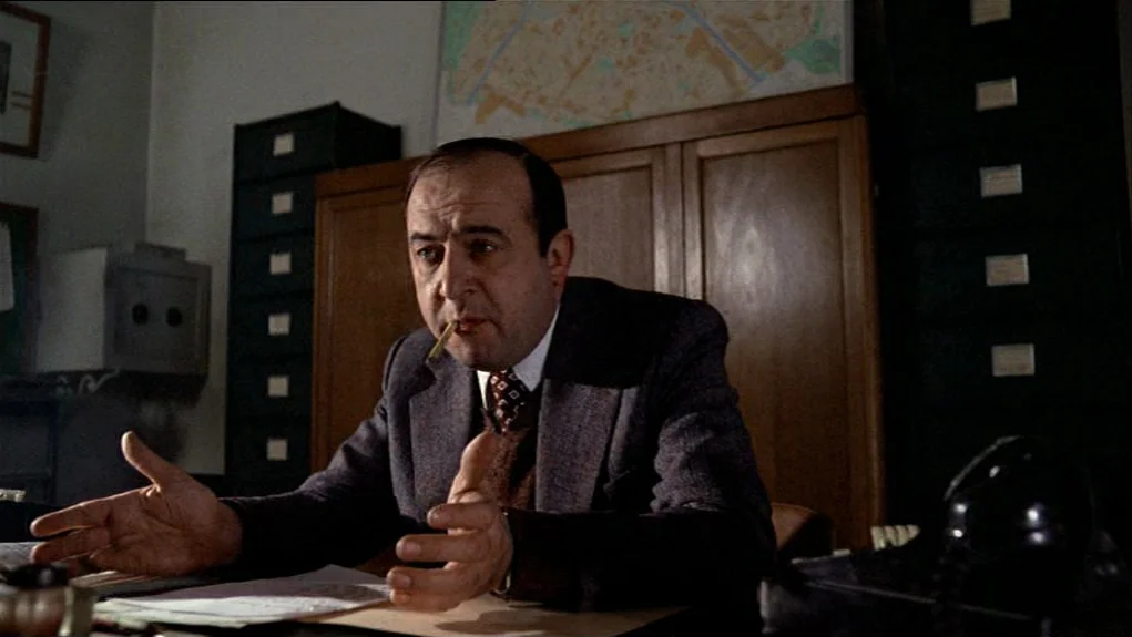 The Tenant (1976) movie still: medium — A bureaucratic interior with wooden cabinets, filing drawers, and a map on the wall. A middle-aged man sits…