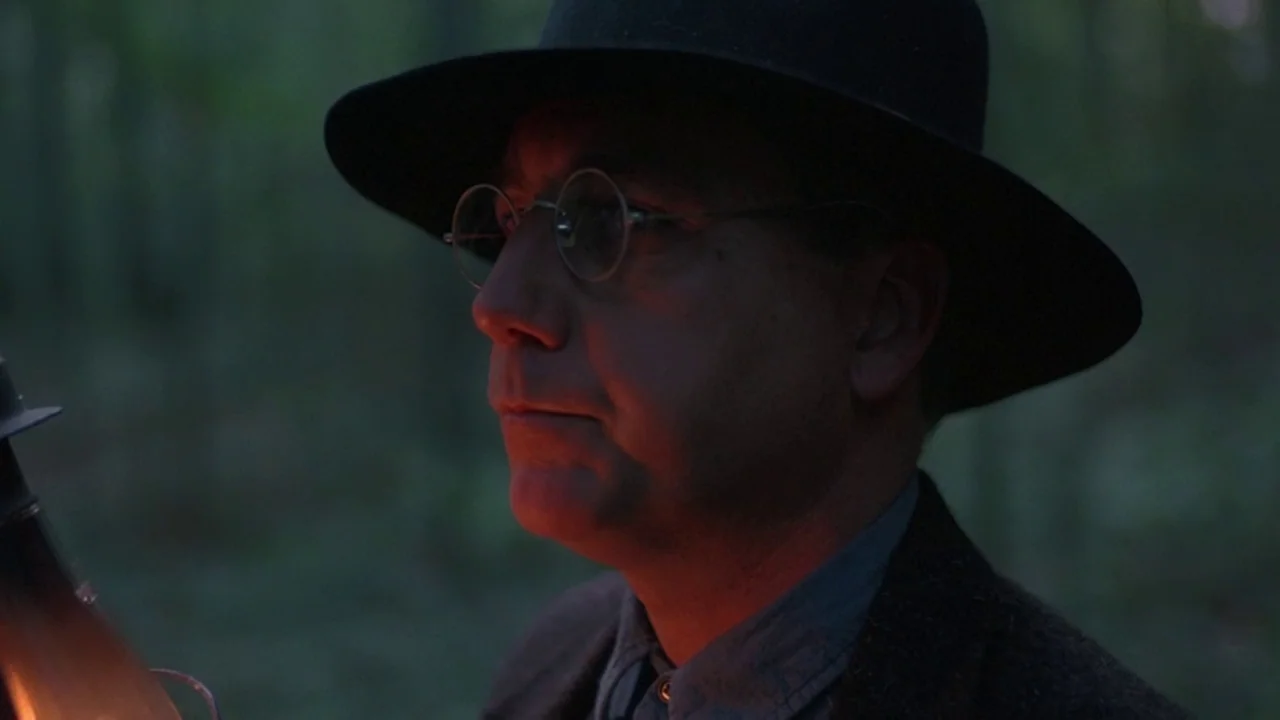 Matewan (1987) movie still: closeup — A dark forest environment at night with dense blurred trees in the background. A man in a hat is lit by a warm…