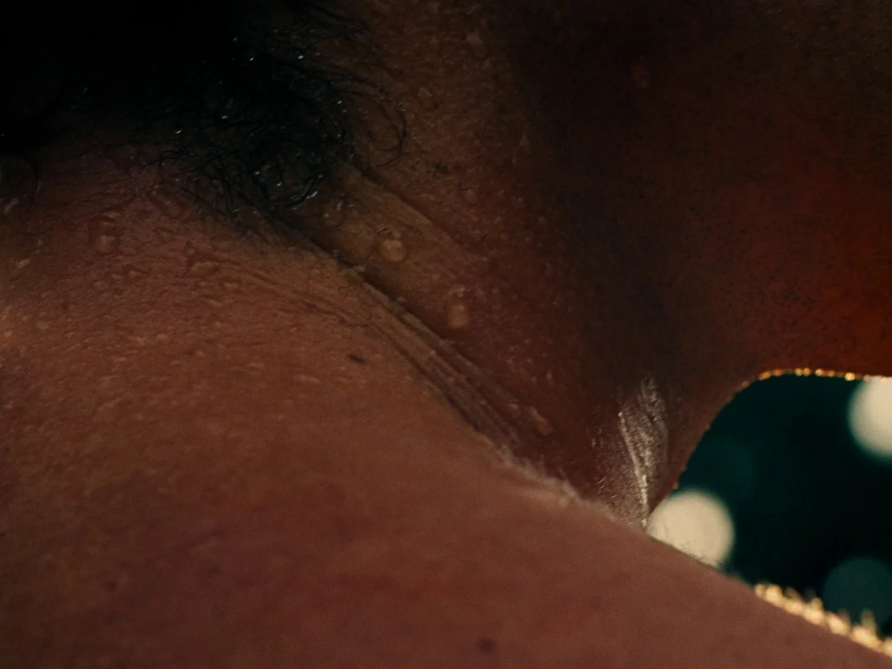 Saltburn (2023) movie still: closeup — A close-up view shows a person's shoulder and neck skin with sweat beads. Sunlight reflects off the skin…