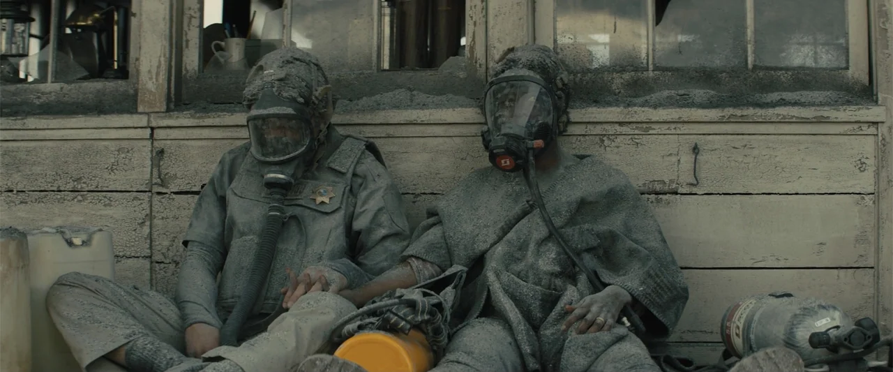 Those Who Wish Me Dead (2021) movie still: medium — Two individuals sit against a weathered, dust-caked wooden wall outside an old building. They…