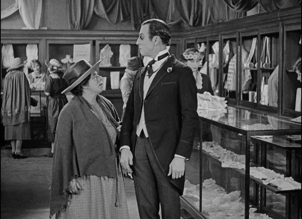Safety Last! (1923) movie still: medium — A 1920s department store interior with glass display cabinets and draped fabric. A woman in a shawl stands…
