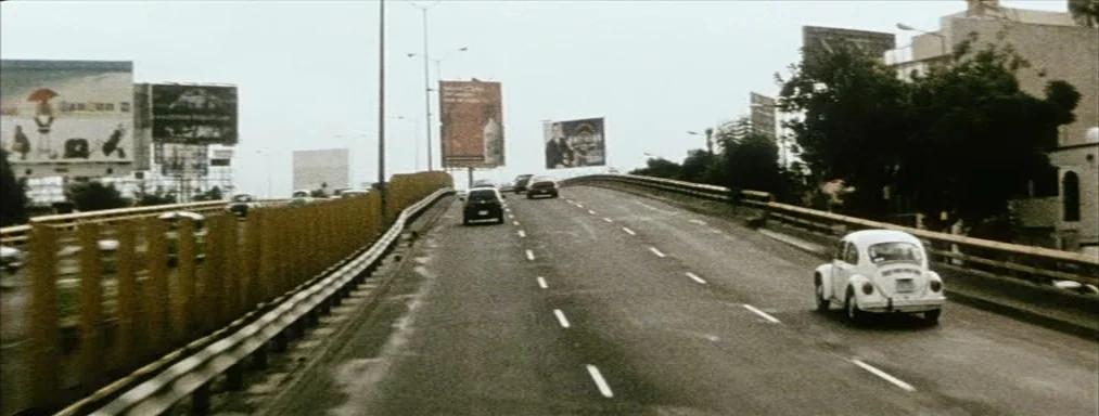 Japón (2002) movie still: wide — A multi-lane highway overpass stretches toward the horizon under a pale, overcast sky. A white vintage Volkswagen…