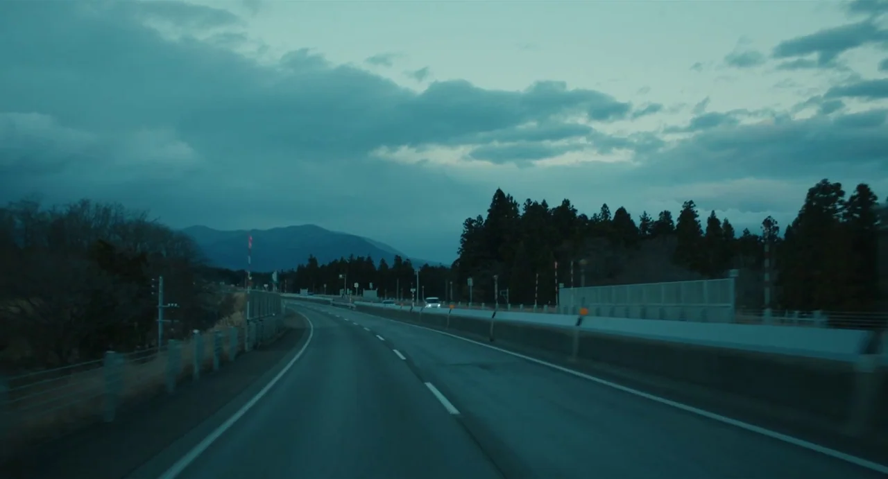 Drive My Car (2021) movie still: establishing — An asphalt road curves through a rural mountainous landscape under a dark overcast sky. Dense…