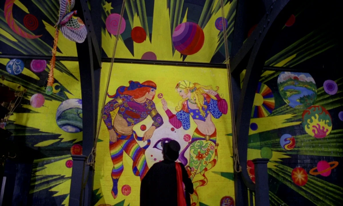 Wonderwall (1968) movie still: medium — An interior space features a large yellow wall covered in a brightly colored psychedelic mural. A man in a…