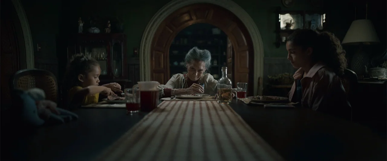 Evil Eye (2020) movie still: medium — A dimly lit dining room features a long table with a central runner fabric. An elderly woman sits in the…