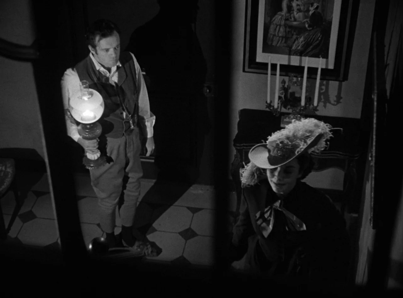Madame Bovary (1949) movie still: medium — A dark, dimly lit foyer features a tiled floor and framed wall art. A man carrying a lantern faces a…