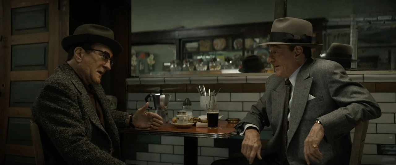 The Alto Knights (2025) movie still: medium — A 1950s diner booth with white tiled walls and a small rectangular table. Two older men wearing suits…