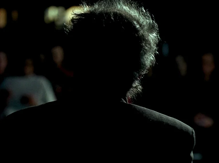 Notre Musique (2004) movie still: closeup — A dark environment at night with an obscured background crowd of blurry figures. A man with textured…