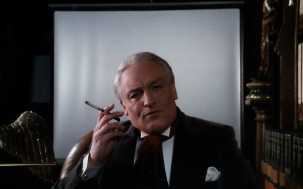 The Rocky Horror Picture Show (1975) movie still: medium — A dimly lit room with books and a projection screen in the background. An elderly man in…