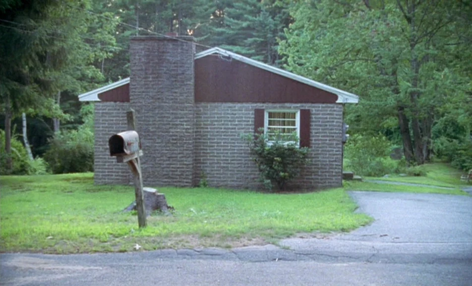 The Brown Bunny (2003) movie still: wide — A single-story brick house sits in a grassy clearing surrounded by dense woods. A weathered mailbox…