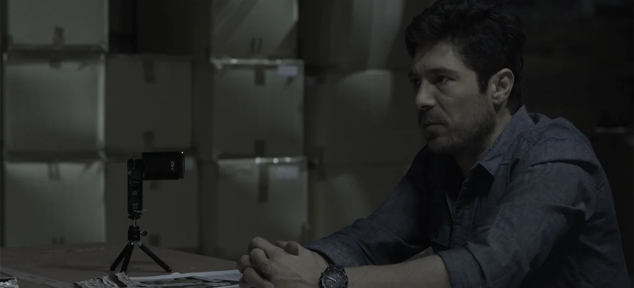 About the Deaths (2016) movie still: medium — A dimly lit warehouse filled with stacks of cardboard boxes in the background. A man sits at a table…
