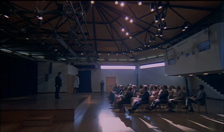 A Clockwork Orange (1971) movie still: wide — A spacious auditorium features a wooden stage and rows of chairs for an audience. A man stands on the…
