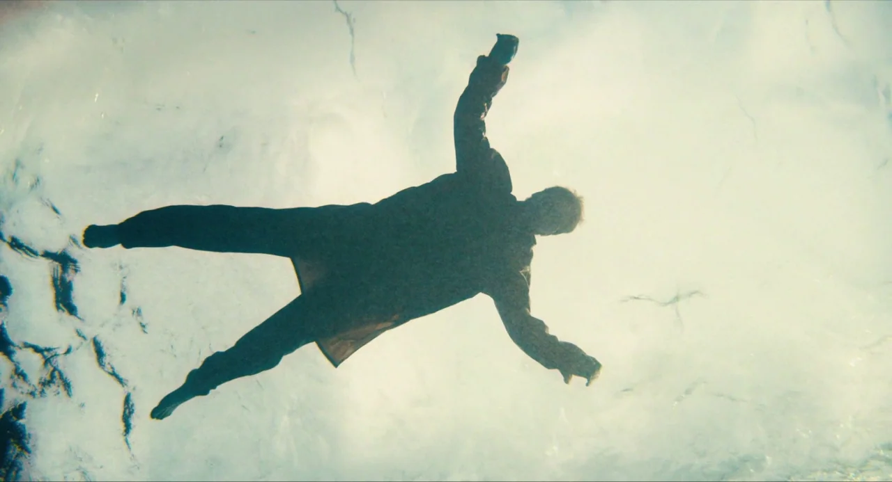 Submergence movie still: fullbody — A dark, silhouetted human figure floats horizontally beneath a bright, rippling water surface. The person is…