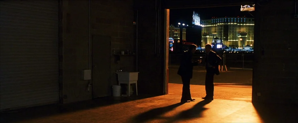 Ocean’s Eleven (2001) movie still: wide — A dark warehouse interior with a concrete wall and utility sink. Two men in dark suits converse while…