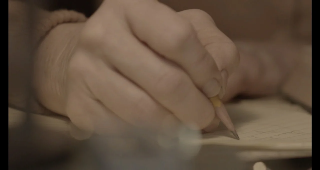 Living With Lincoln (2015) movie still: closeup — A wooden desk surface holds a sheet of lined paper with handwritten text. A hand holds a yellow…