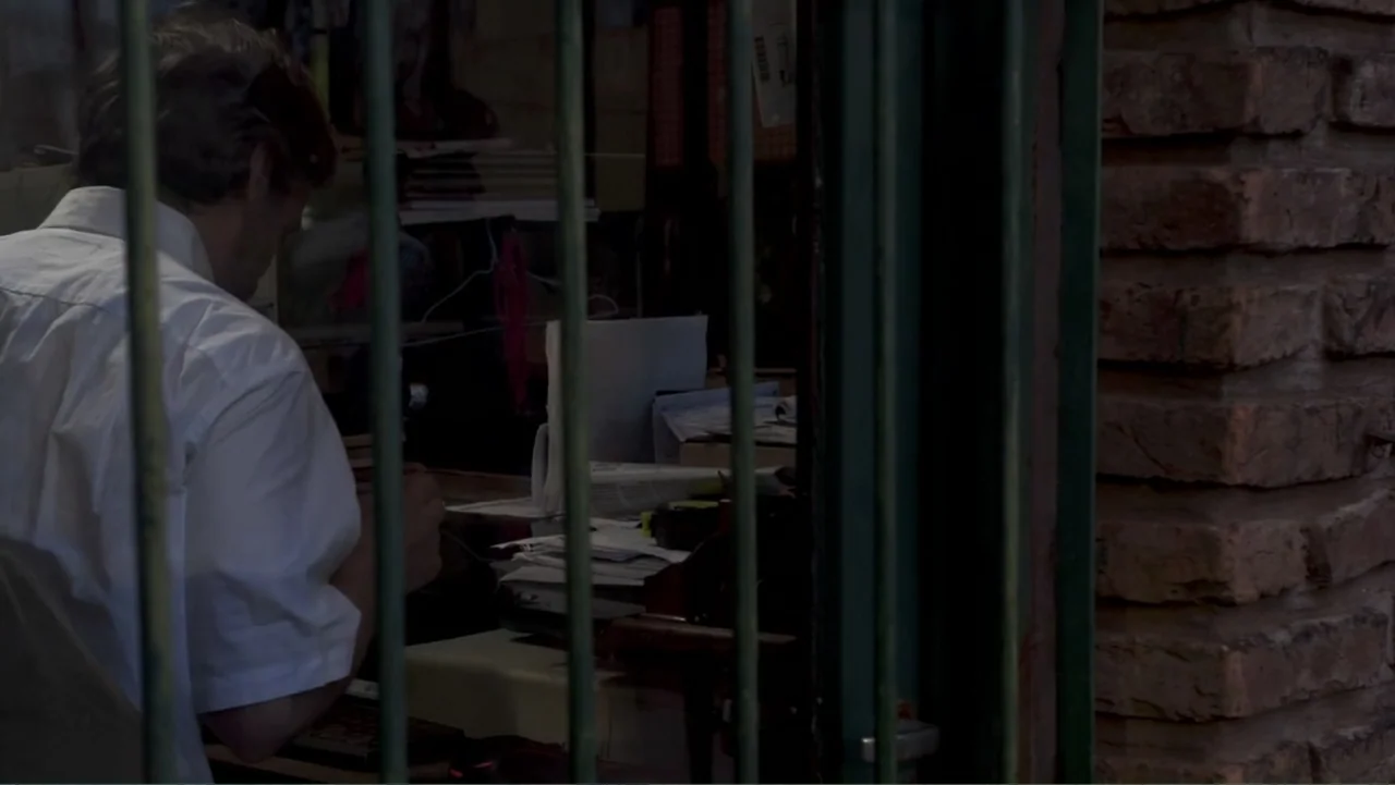 Smog in Your Heart (2022) movie still: medium — An interior office space seen through vertical metal window bars features a messy desk. A man in a…