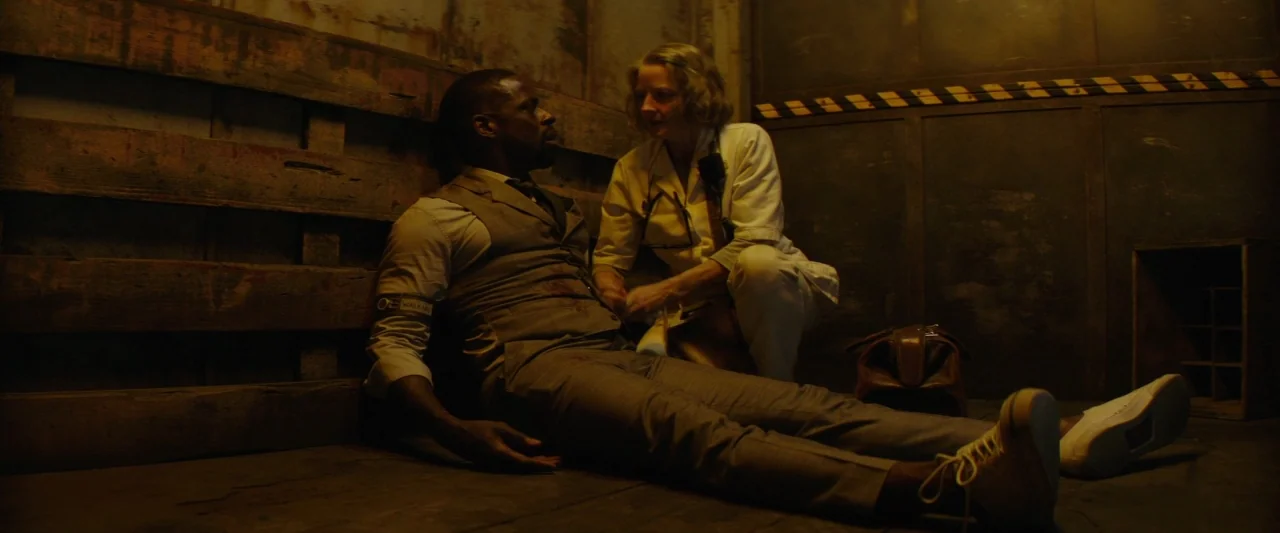 Hotel Artemis (2018) movie still: medium — Two individuals sit on the floor of a dark room with horizontal wooden wall slats. A medical professional…