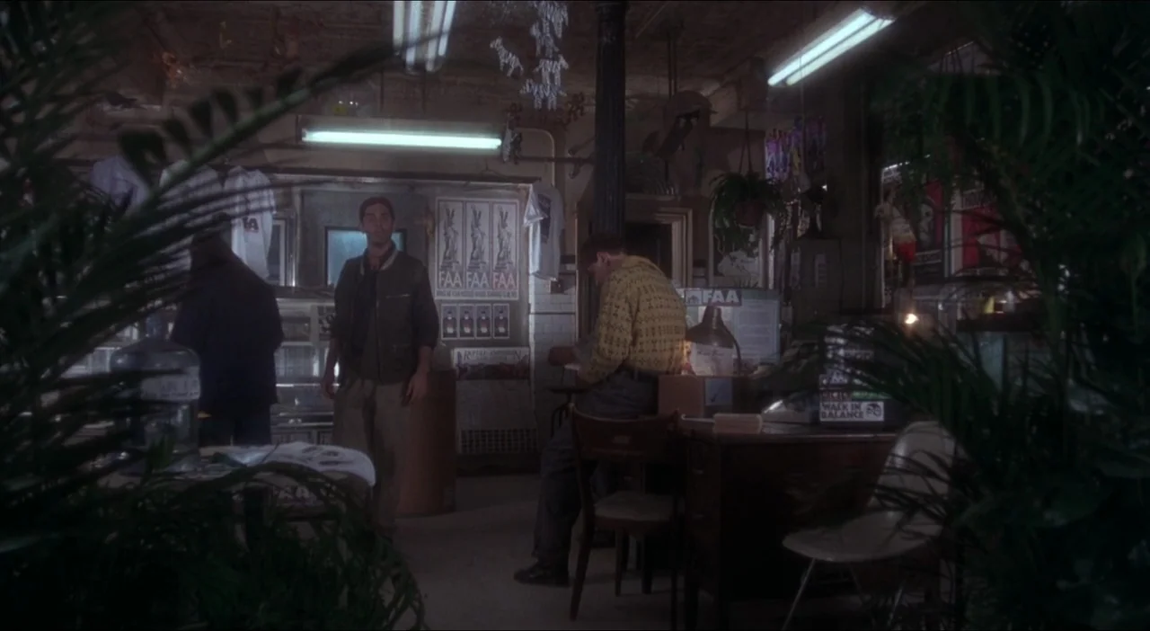 Twelve Monkeys (1995) movie still: wide — A cluttered shop interior features low ceilings, hanging decorations, and dim overhead fluorescent…