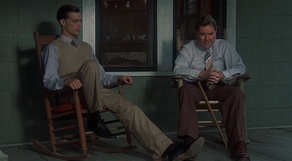 Thieves Like Us (1974) movie still: fullbody — A green wooden porch serves as the setting for two men relaxing. Both men sit in wooden rocking…