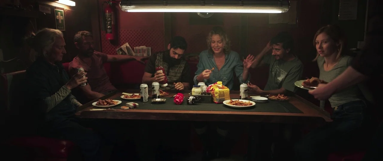 Sea Fever (2019) movie still: wide — A dimly lit restaurant booth with red vinyl seating accommodates six people around a table. The group eats fast…