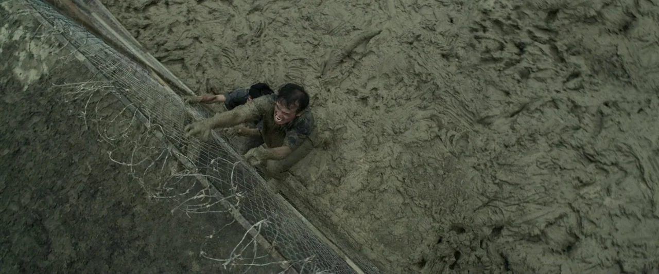 The Raid 2 (2014) movie still: wide — A desolate expanse of deep churned mud borders a tall chain-link and barbed-wire fence. Two men covered in mud…