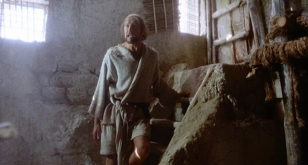 Life of Brian (1979) movie still: medium — A rustic interior with thick stone walls and a set of crude earthen stairs. A disheveled man in simple…