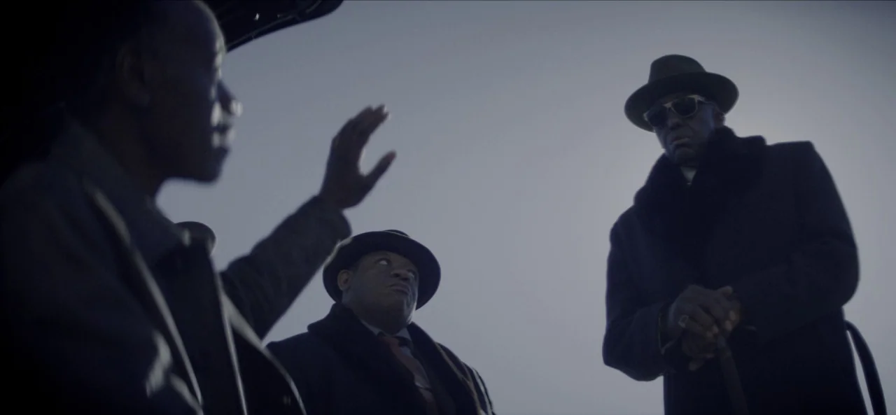 No Sudden Move (2021) movie still: medium — A group of three men stand together under an overcast, grey sky. One man is gesturing with his hand…