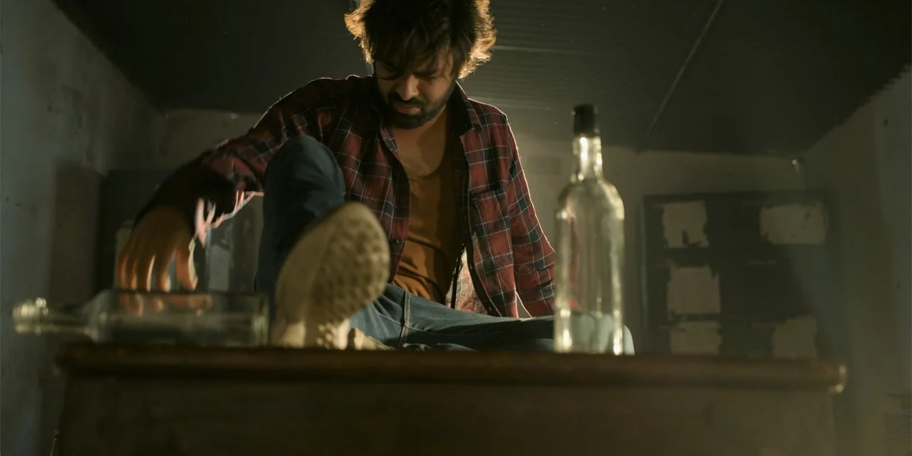 Dry Day (2023) movie still: medium — A dark, sparsely furnished interior with muted walls and a heavy wooden table. A man sits atop the table in a…