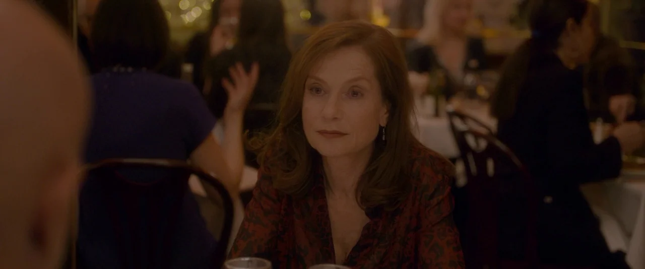 Elle (2016) movie still: medium — A crowded restaurant with multiple tables and blurred patrons in the dim background. A middle-aged woman sits at a…