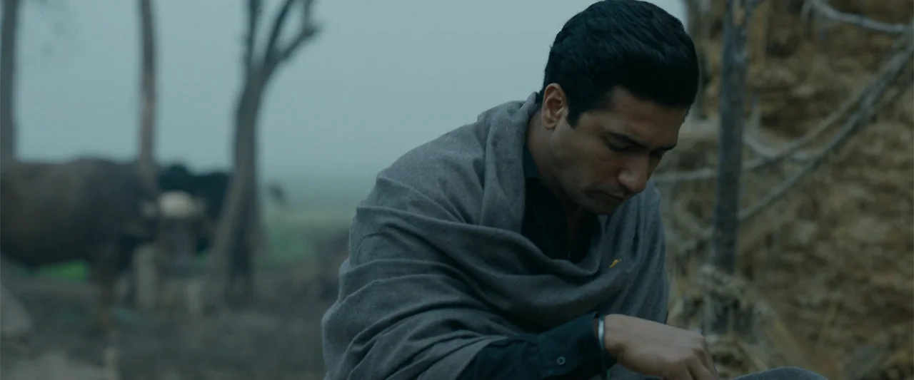 Sardar Udham (2021) movie still: medium — An outdoor rural area features sparse trees and a hazy, overcast horizon. A man in a dark shirt and grey…