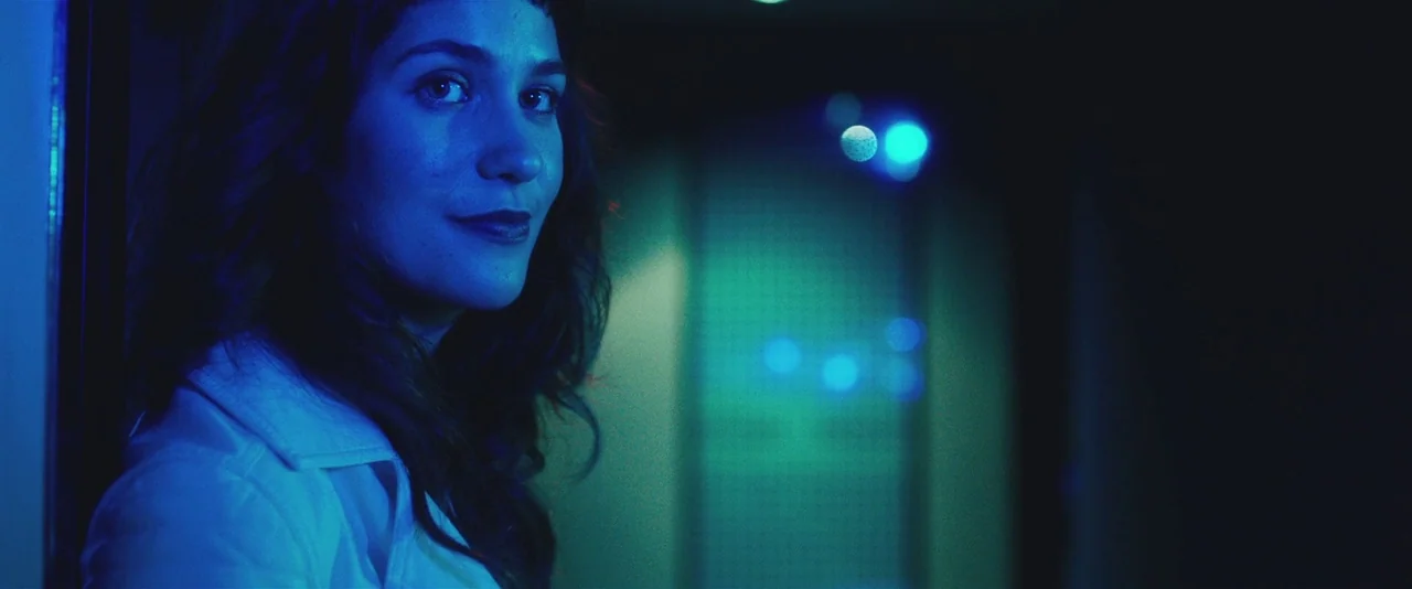 Gemini (2017) movie still: closeup — A dark, blurred corridor is illuminated by distant blue and cyan neon lights. A young woman stands in the…