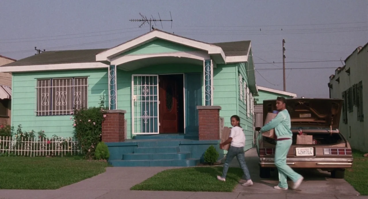 Boyz In The Hood (1991) movie still: wide — A suburban mint-green home features a white security gate and dark brick pillars. Two females carry…