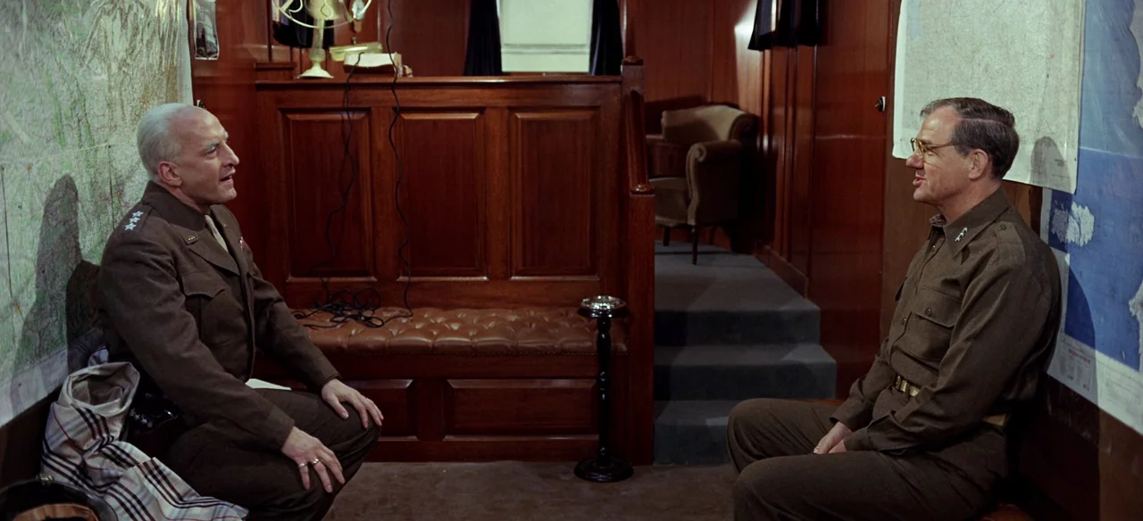 Patton (1970) movie still: medium — A wood-paneled room contains maps on walls, a leather bench, and steps leading upward. Two military officers in…
