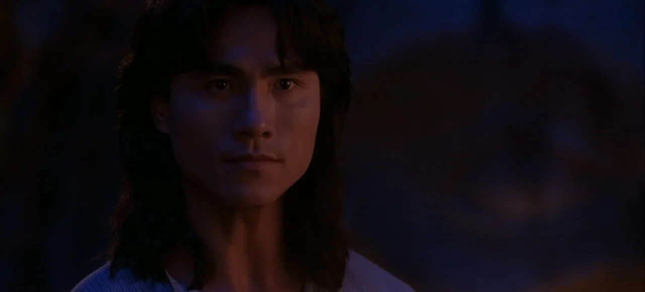 Mortal Kombat (2021) movie still: closeup — A man stands in a dimly lit, indistinct interior space with deep shadows. The subject gazes towards the…