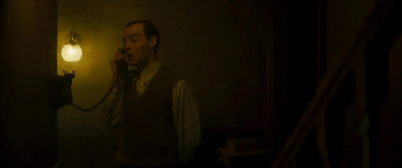 Earwig (2022) movie still: medium — A dark, shadows-heavy residential interior with a wall-mounted telephone and a staircase railing. A man in a…